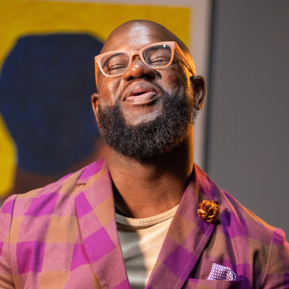 Portrait of John Lumpkin II wearing glasses and a suit, looking upward against a warm-toned patterned background.